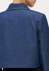 Blå denimjakke med kort kutt, enkelt nothals, og hvite sømmer langs sømmene. Glatt tekstur.