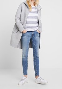 Světle šedá oversize bunda s kapucí přes bílou a šedou pruhovanou tričko, doplněná modrými slim džíny a bílými teniskami.