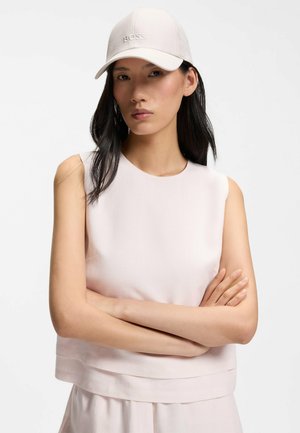 Nainen, jolla on pitkät tummat hiukset, vaalean värinen hihaton paita ja valkoinen lippalakki, jossa on "BOSS"-logo, kädet ristissä, neutraali ilme valkoista taustaa vasten.