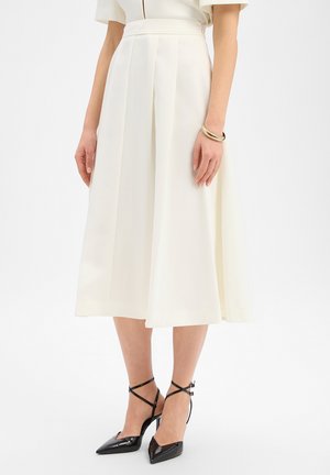 Frau trägt weiße Culottes mit hohem Bund und weitem Bein sowie schwarze spitze Pumps mit Knöchelriemen, steht mit locker hängenden Armen an den Seiten.