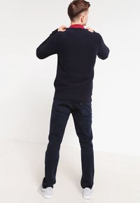 Marinblå stickad tröja med ett texturerat mönster och ribbade mudd, bärs över en röd skjorta med krage; matchad med mörka denimjeans och vita sneakers.