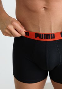 Boxeri negri din material elastic, cu un brâu elastic roșu îndrăzneț ce prezintă inscripția "PUMA" în litere negre. Textură netedă și croială ajustată.