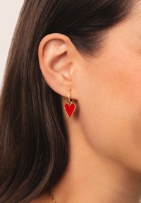 Goldfarbene Creolen mit einem roten herzförmigen Anhänger. Die Ohrringe sind schlank mit einer glatten Emaille-Oberfläche und einem glänzenden Metallelement.