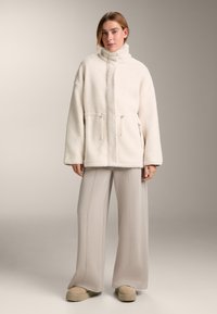 Giacca crema in finto shearling con colletto alto, cerniere frontali e tasche laterali. Abbinata a pantaloni beige chiaro ampi e scarpe con plateau.