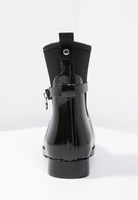 Bottine noire brillante avec tige en tissu, languette à l'arrière, sangle décorative avec boucle et petit talon carré, posée sur une surface blanche.