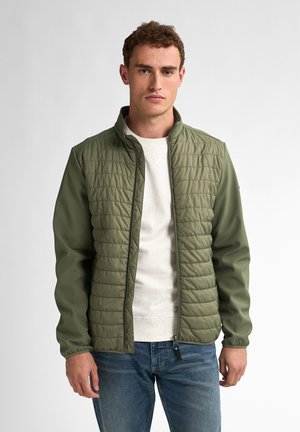 Chaqueta acolchada verde con cuello alto, textura suave, cierre con cremallera y mangas largas, llevada sobre un suéter de color claro y jeans azules.