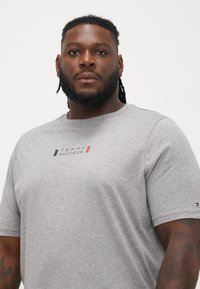 Szara koszulka z krótkim rękawem wykonana z bawełny, z małym czerwonym i granatowym logo z napisem "TOMMY HILFIGER" na klatce piersiowej oraz naszywką logo na rękawie.