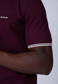 Polo shirt bordeaux en tissu texturé. Présente un petit graphique d'animal blanc et des poignets de manches rayés en blanc et marron.
