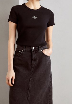 Kvinde iført en tætsiddende sort top med korte ærmer, et lille geometrisk logo og en høj taljet mørk denimnederdel, med hånden i lommen.