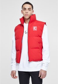 Rote Steppweste mit hohem Kragen und Reißverschluss. Mit einem Logo auf der Brust und über einem weißen Langarmshirt getragen.