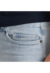 Himmelblaue Denim-Jeans mit einer kleinen Vordertasche und einem silbernen Metallknopf-Detail. Der Stoff wirkt strukturiert mit einem leichten Verblassen.