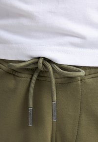 Un pantalon de survêtement vert olive avec un cordon en tissu doté d'embouts en métal, associé à un t-shirt en coton blanc, montrant les textures et les détails de la ceinture.