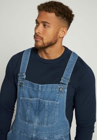 Salopette in denim con bretelle regolabili, tasca sul petto e colore blu sbiadito, abbinata a una camicia a maniche lunghe blu navy sotto.