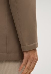 Braune Jacke mit strukturierter Oberfläche, ausgestattet mit umgeschlagenem Ärmel und kontrastierender Nahtgestaltung. Hand sichtbar, die auf einer Cordoberfläche ruht.