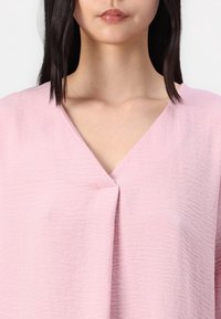 Top rosa realizzato in tessuto testurizzato, con design a V e una piega drappeggiata sul davanti, creando una silhouette morbida e rilassata.