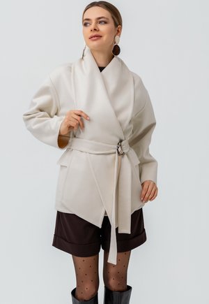 Beżowa kurtka typu wrap z szerokimi rękawami i paskiem w talii z okrągłą metalową klamrą. Noszona na ciemnych szortach i rajstopach w kropki.