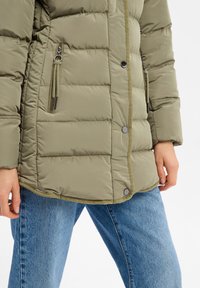 Zielona oliwkowa kurtka puchowa o pikowanym wzorze, z zamykanymi kieszeniami i zatrzaskami, w połączeniu z jasnoniebieskimi dżinsami.