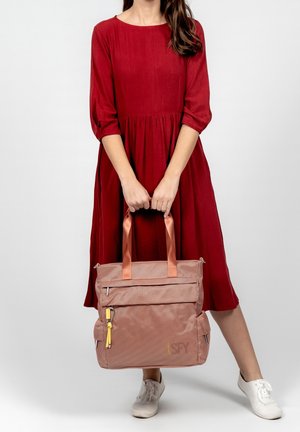 Borsa a tracolla rosa con texture, due manici, tasca frontale con zip e zip gialla, tenuta da una persona in un abito rosso e sneakers bianche.