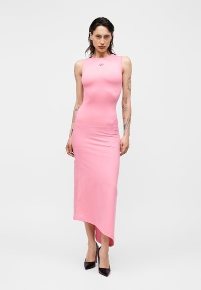 Λεπτή γυναίκα με τατουάζ φοράει ασύμμετρο ροζ μίντι φόρεμα χωρίς μανίκια και μαύρες ψηλοτάκουνες, στέκεται μπροστά σε απλό φωτεινό φόντο.