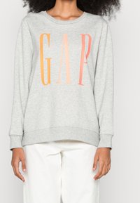 Sudadera gris claro con cuello redondo, que presenta grandes letras en degradado que dicen "GAP" en naranja y rosa. Puños y dobladillo de canalé.