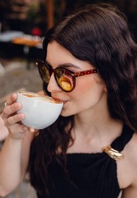 Hnědé želvovinové sluneční brýle, velký kulatý tvar, se zlatými akcenty. Bílý keramický hrnek držený v levé ruce, naplněný kávou, hladký povrch.