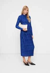 Mėlyna ilga rankovė su tekstūruotu raštu, prigludusi bodice ir tiesi siluetas. Modelis laiko baltą delninę ir dėvi juodus rankovinius batelius.