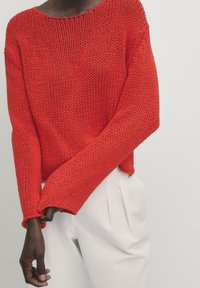 Pull en maille rouge à coupe ample, manches larges et col rond, associé à un pantalon blanc plissé. Tissu légèrement texturé.