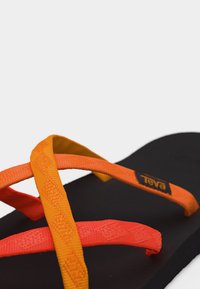 Πέδιλο με μαύρη σόλα και σταυρωτά πορτοκαλί και κόκκινα υφασμάτινα λουριά, με ένα μικρό λογότυπο Teva σε ένα από τα πορτοκαλί λουριά.
