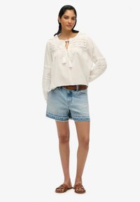 Blusa leggera bianca con dettagli in pizzo e lacci a nappina; abbinata a pantaloni corti in denim azzurro con orlo sfrangiato; sandali in pelle marrone.