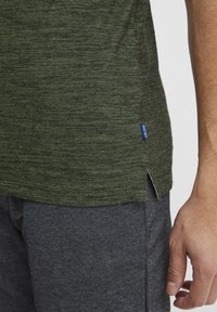 Olivengr�øn, tekstureret kortærmet skjorte med sideslids og et lille blåt mærkat, parret med mørkegrå teksturerede shorts.