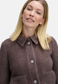 Brun uldjakke med krave, frontlukning med knapper, to lommer og en tekstureret overflade. Har store grå knapper.