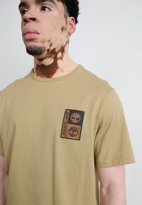 Khaki t-shirt met korte mouwen, gemaakt van katoen, met een zwart Timberland-logo op de linkerborst. Soepele textuur en een comfortabele pasvorm.