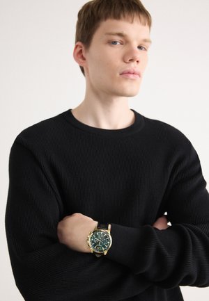 Zwarte ribbeltrui met een ronde hals; model draagt een chronograaf horloge in goud en groen met een zwarte leren band.