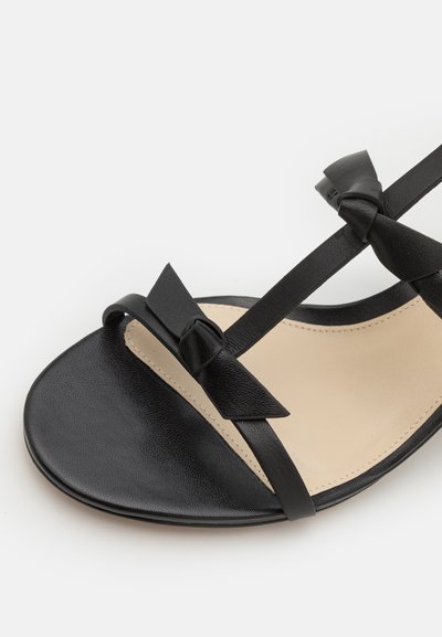Svart sandal med ett slätt läderöverdrag med rosettdekorationer, en platt sula och minimala remmar, placerad på en beige innersula.