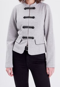 Veste grise sur mesure avec col montant, ornée de fermetures à bascule noires décoratives et poches latérales. Texture lisse et coupe ajustée.