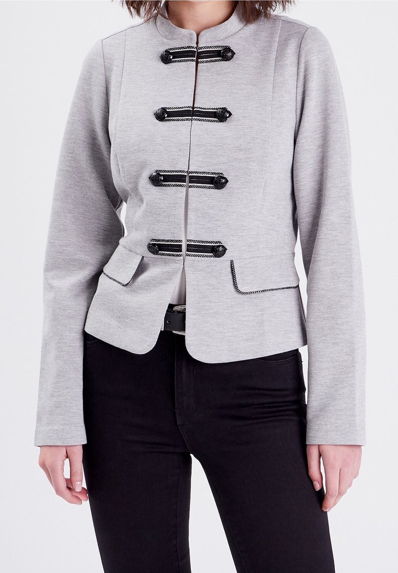 Veste grise sur mesure avec col montant, ornée de fermetures à bascule noires décoratives et poches latérales. Texture lisse et coupe ajustée.