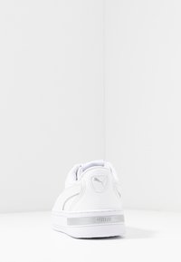 Bílé tenisky s texturovaným koženým svrškem a polstrovaným límcem. Mají stříbrné logo Puma na patě a bílou gumovou podrážku. Minimalistický design.