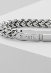 Stříbrný kovový náramek s proplétanými řetězovými články a závěsným zapínáním, na kterém je vyryto "BREIL." Hladký a leštěný povrch.