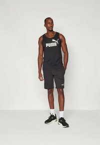 Άνδρας που φοράει μαύρη μπλούζα από Puma, μαύρες βερμούδες, λευκές κάλτσες και μαύρα αθλητικά παπούτσια, στέκεται μπροστά σε απλή ανοιχτή φόντο.
