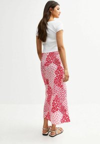 Top corto a maniche corte bianco a coste abbinato a una gonna midi trasparente con motivo floreale rosa e rossa, sandali piatti con cinturini e lunghi capelli scuri.