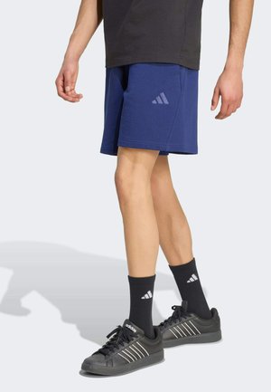 Osoba nosząca niebieskie szorty Adidas, czarne skarpetki Adidas i czarne buty sportowe Adidas, stojąca z widocznymi nogami na jasnym tle.