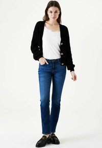 Cárdigan negro de punto con mangas oversize y botones brillantes, sobre un camisón blanco y combinado con jeans azules y mocasines negros.