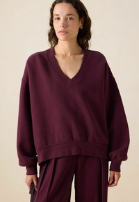 Sweatshirt bordeaux surdimensionné avec un col en V et des manches longues froncées. Texture douce avec une coupe décontractée et un détail de bord côtelé.