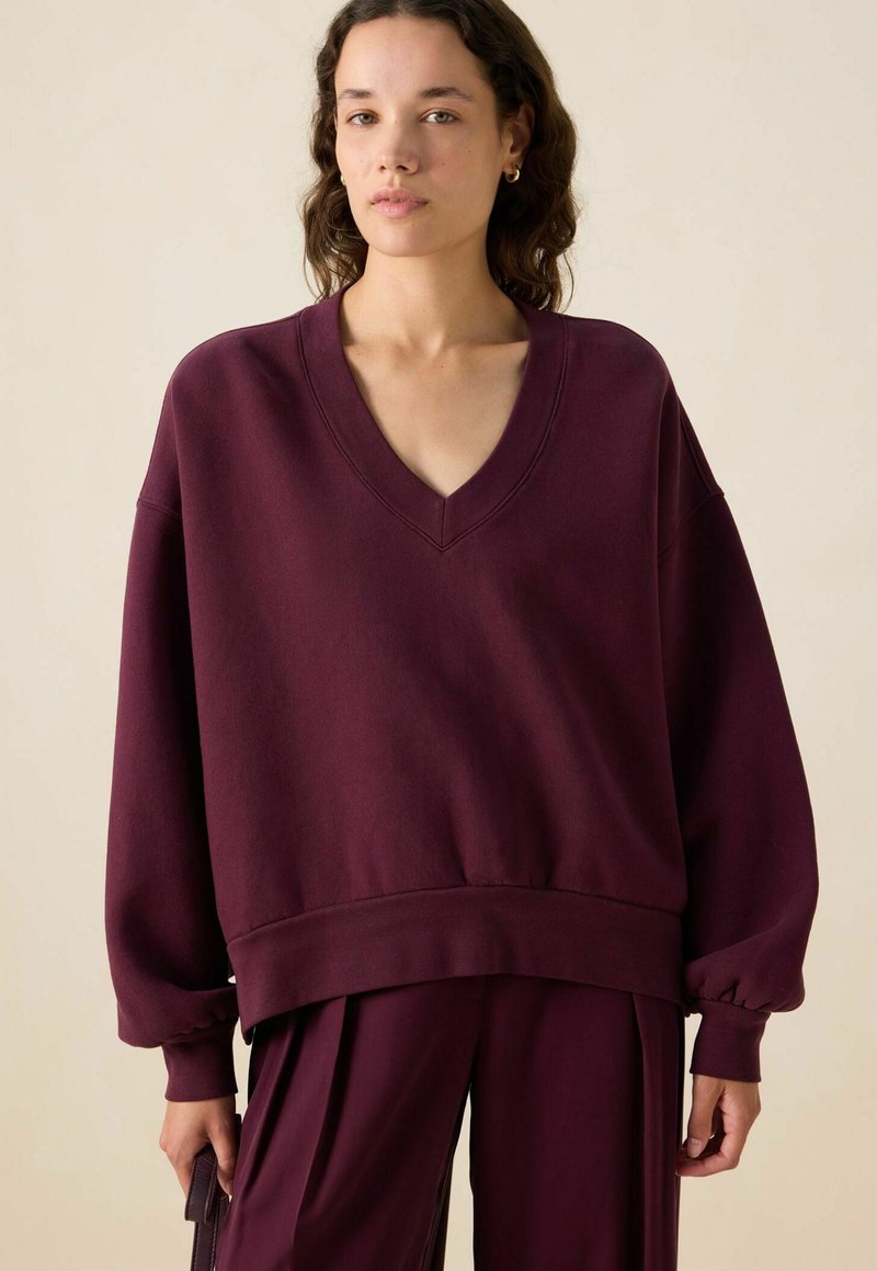 Sweatshirt bordeaux surdimensionné avec un col en V et des manches longues froncées. Texture douce avec une coupe décontractée et un détail de bord côtelé.