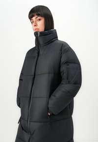 Woman with short dark hair wearing a black puffer jacket with high collar and zippered pockets, hands in side pockets, neutral background.