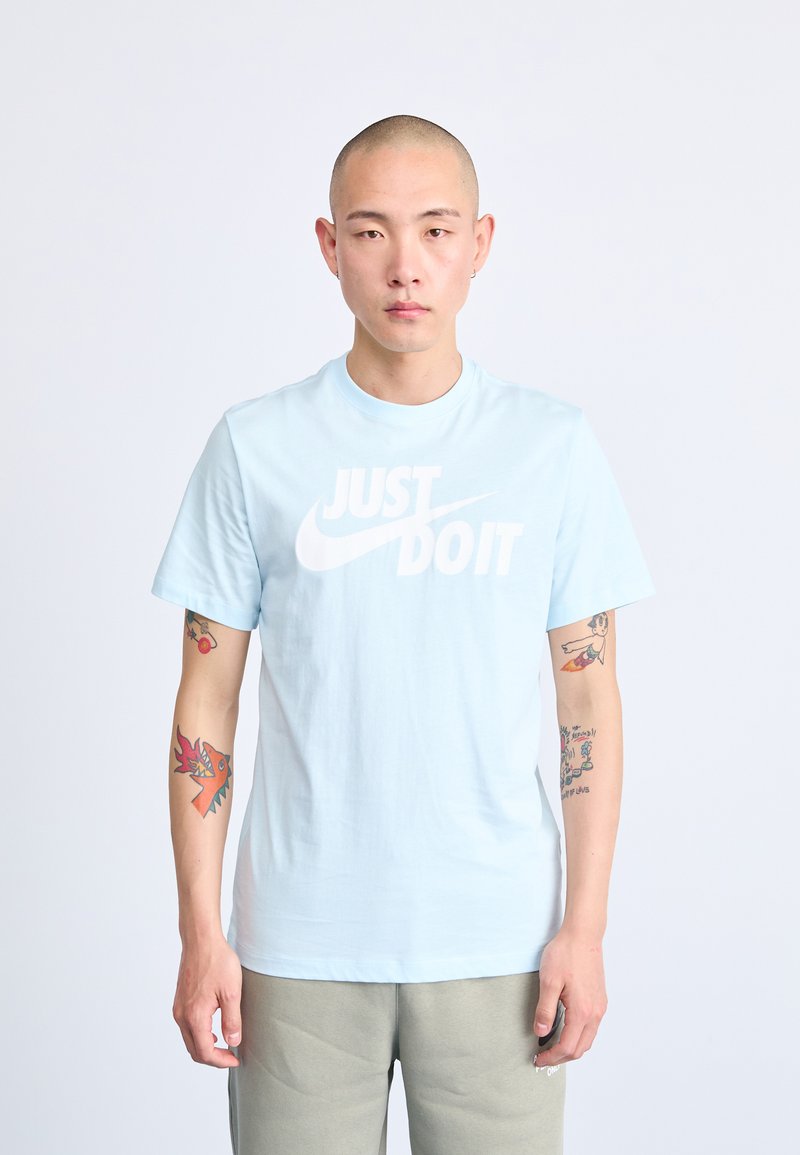 Šviesiai mėlyna medvilninė marškinėliai su trumpomis rankovėmis, su dideliu baltu „JUST DO IT“ grafiniu dizainu. Modelio rankos padengtos matomomis tatuiruotėmis.