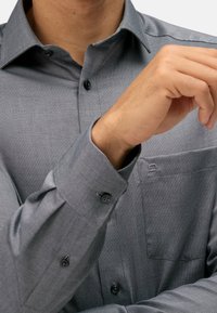Camicia grigia con bottoni caratterizzata da un tessuto texturizzato, colletto a punta e una tasca sul petto con un piccolo logo vicino al colletto.