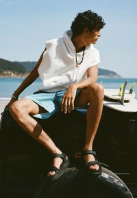 Černé sandály se straps, světle modré �šortky, bílé tílko bez rukávů. Sedí na vozidle, v pozadí surfovací prkna, viditelné moře.