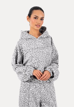 Kvinde iført grå og hvid leopardmønstret hættetrøje og matchende bukser, stående med hænderne foldet foran.