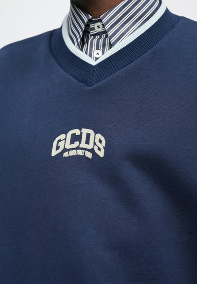 Sweater azul-marinho de decote em V, feito de um tecido suave, apresentando um logótipo bordado em branco "GCDS MILAN ITALY 1983". Debaixo, uma camisa às riscas é visível.